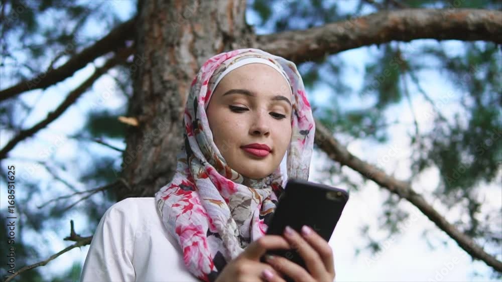 A young Muslim woman in a traditional headscarf holding a smartphone in his hands, a lady communicating with friends on her mobile phone in the park, she sits outside near a tall tree A young Muslim woman in a traditional headscarf holding a smartphone in his hands, a lady communicating with friends on her mobile phone in the park, she sits outside near a tall tree
