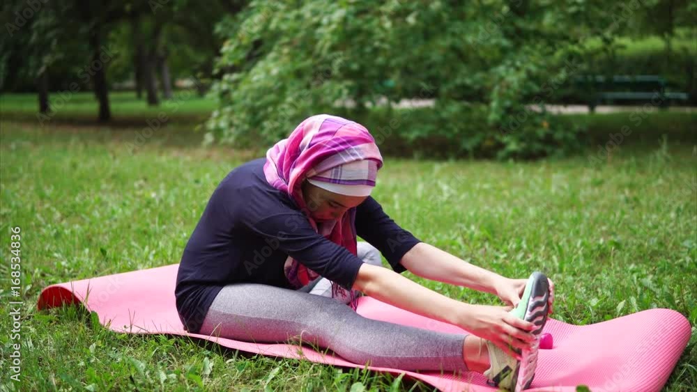 Young muslim woman in a scarf that covers her hair doing exercises for stretching the muscles of the hip, the lady is sitting on the rug for yoga Young muslim woman in a scarf that covers her hair doing exercises for stretching the muscles of the hip, the lady is sitting on the rug for yoga