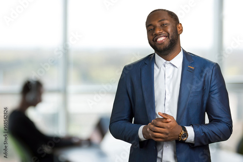 Smiling black businessman in office. Afro american man in suit in good mood. Handsome black manager of company smiling on blurred background.
