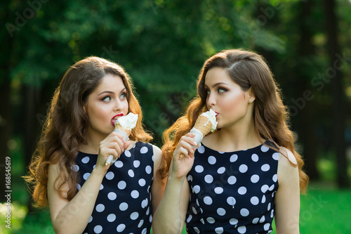 Two girlfriends sister twins of the same appearance with makeup and hairdo in the summer park eating ice cream