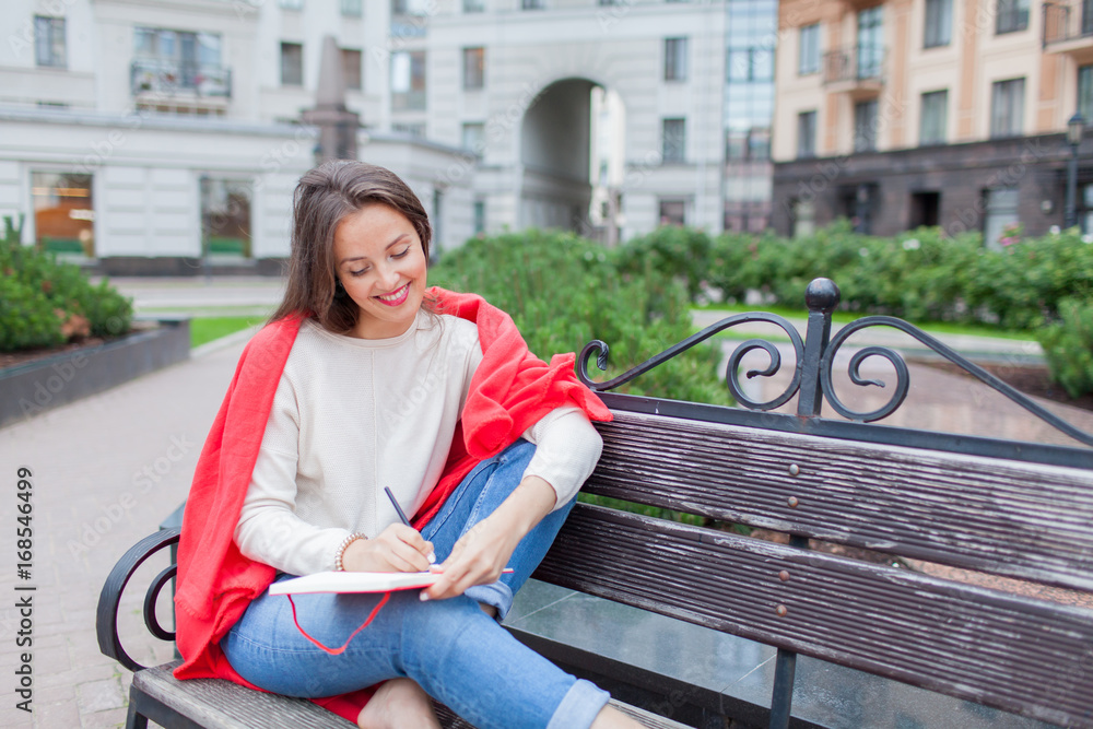 Attractive girl sitting on a bench with bare feet, covered with a red blanket, in the new residential area and writes his thoughts into a red notebook. The urban background Attractive girl sitting on a bench with bare feet, covered with a red blanket, in the new residential area and writes his thoughts into a red notebook. The urban background