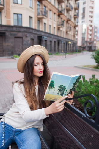 A young beautiful woman in an elegant hat sits on a bench in a new residential neighborhood and reads a paper book. She flips through the pages. Urban background