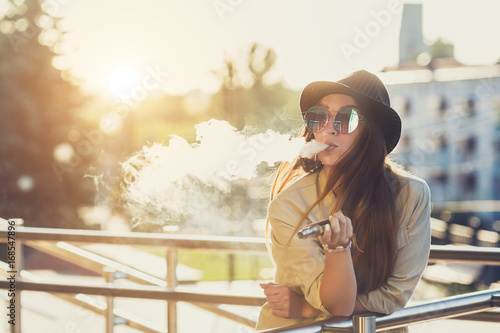 Pretty young hipster woman in black hat vape ecig, vaping device at the sunset. Toned image. photo