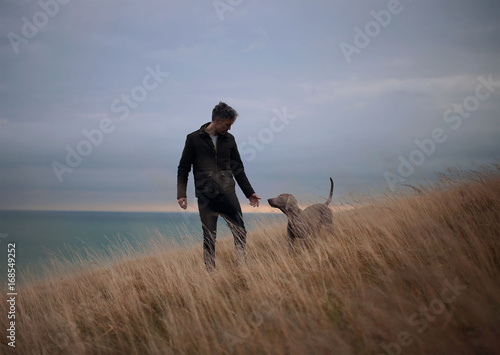 Man with dog walking on grassy landscape against sky