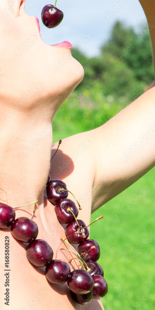 young woman in beads made of fresh cherry holding cherry on open mouth with summer green forest background