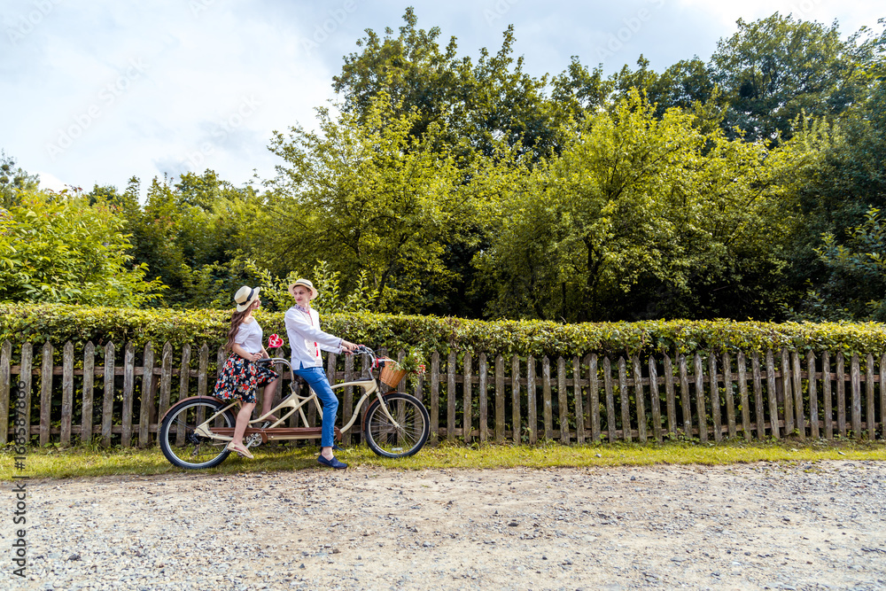 Fototapeta premium Young couple riding a bike tandem in the park. Against the background of the fence from the tree