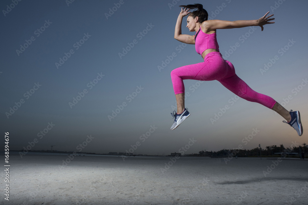 A side view of a strong athletic, beautiful female sprinter as she does a running jumping with an early morning sunrise and dark silhouettes of homes wearing a tight pink fitness outfit  