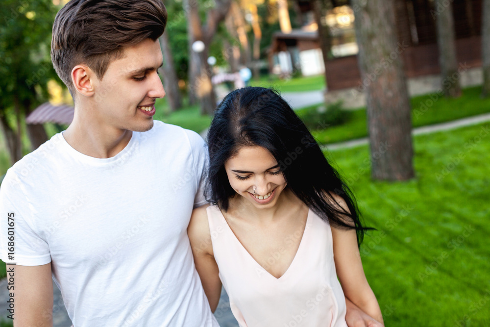 The guy hugs the waist of the girl with a shy smile while walking in nature. Beautiful young couple during a romantic date. The guy hugs the waist of the girl with a shy smile while walking in nature. Beautiful young couple during a romantic date.