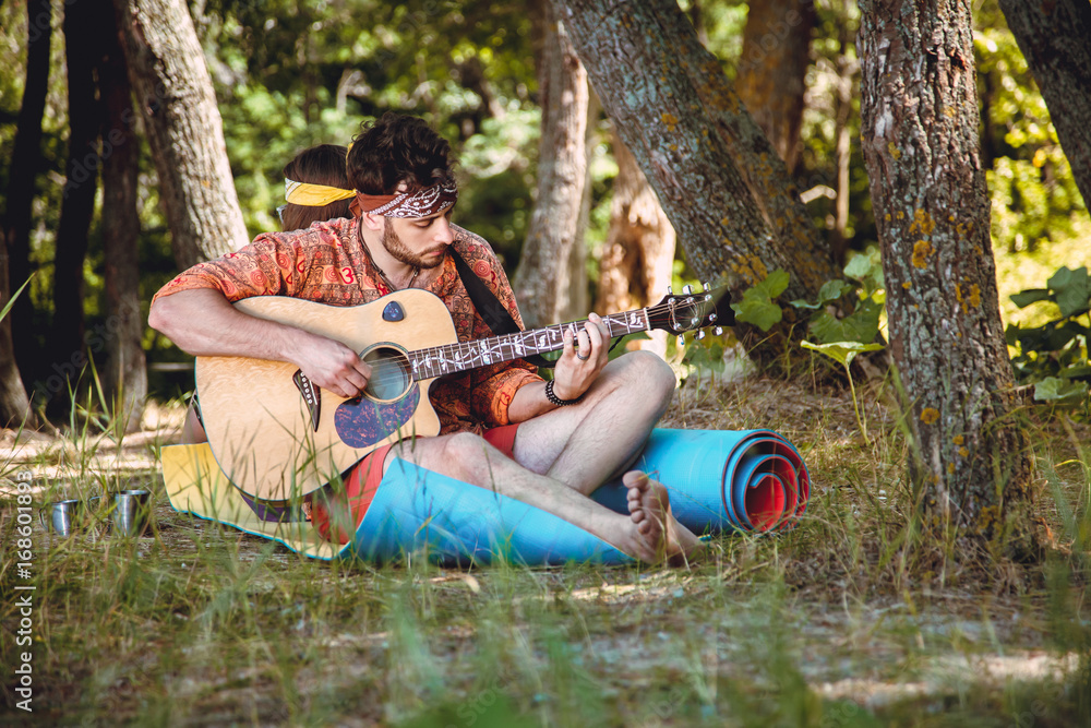 Fototapeta premium Beautiful young couple in love in hippie style rest on the nature. A man plays the guitar and the girl plays the drum