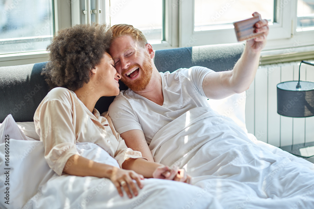 Fototapeta premium Young couple making selfie while lying in bed