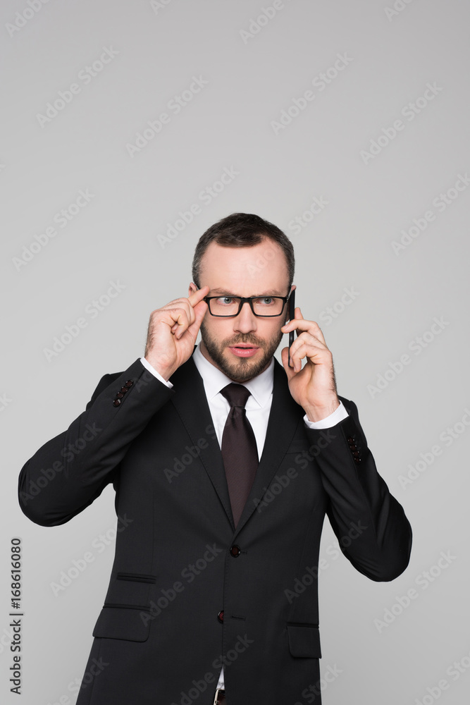 Half-length shot of young businessman in a suit talking on the phone and touching his glasses with concerned look on his face Half-length shot of young businessman in a suit talking on the phone and touching his glasses with concerned look on his face