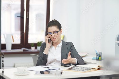 Hard-working entrepreneur in eyeglasses conducting telephone negotiations with business partner while sitting desk in modern office