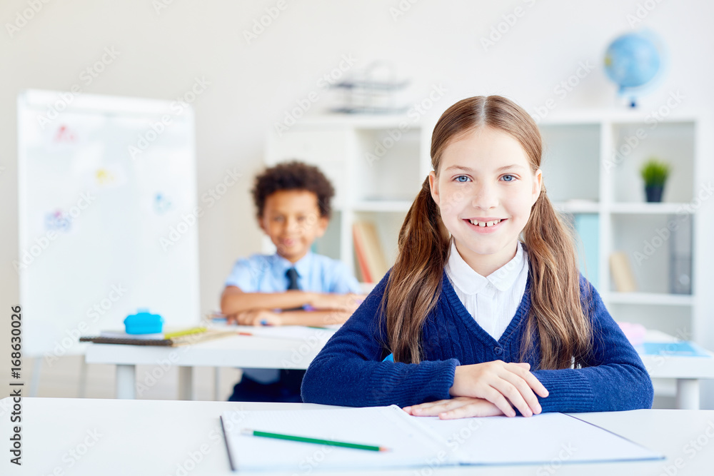 Pretty scholar sitting by desk with classmate on background Pretty scholar sitting by desk with classmate on background
