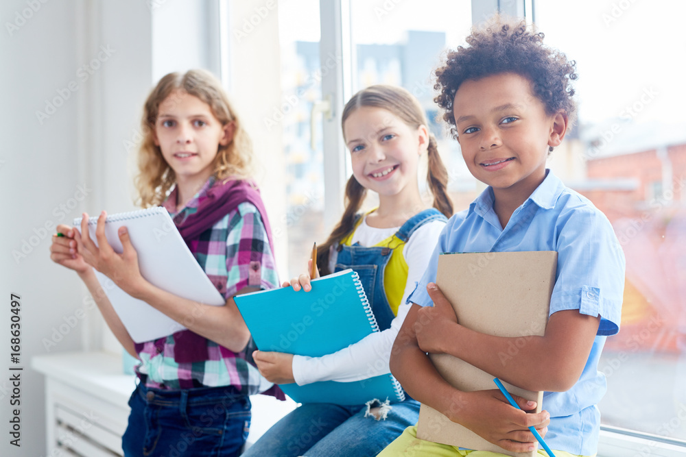 Group of kids with copybooks and pencils sitting by classroom window Group of kids with copybooks and pencils sitting by classroom window