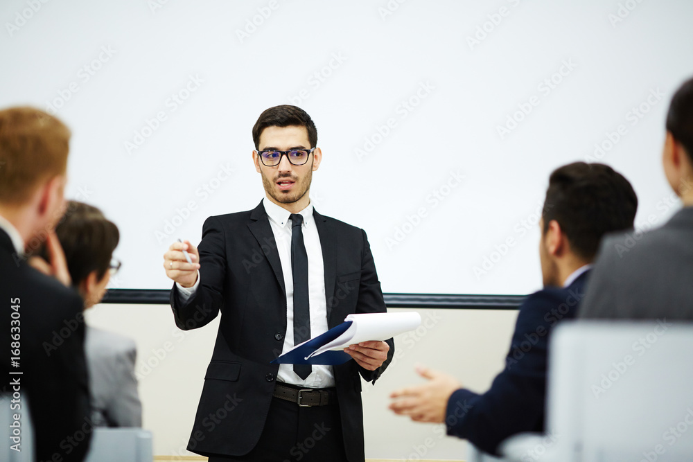 Confident young speaker in stylish suit exchanging ideas with audience while having business conference Confident young speaker in stylish suit exchanging ideas with audience while having business conference