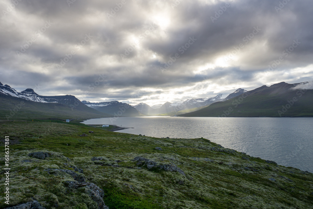  Iceland - Sun breaking through clouds at fjord with mountains and green moss covered landscape