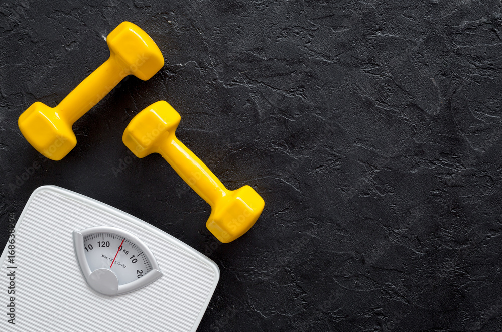 Fototapeta premium Fitness for losing weight. Bathroom scale and dumbbell on black background top view
