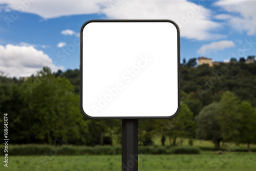 Square white sign with rounded corners. In the background a park and a hill with houses. The background is blurry.