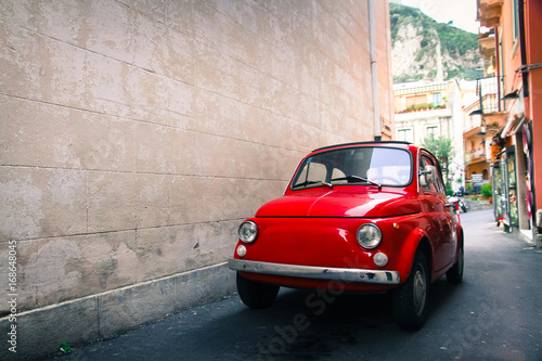 Red old well-preserved vintage Italian classic car parked in a small alley in an Italian Sicilian city with a large empty stone wall in the left side of the frame.