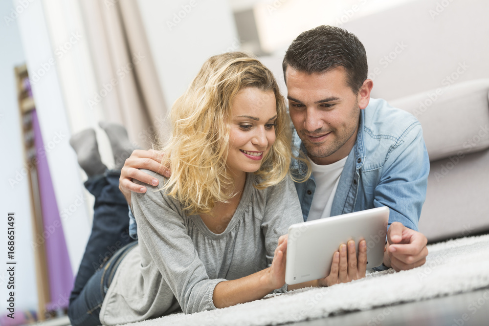 Happy young couple layed on the carpet of their living room and using a tablet Happy young couple layed on the carpet of their living room and using a tablet
