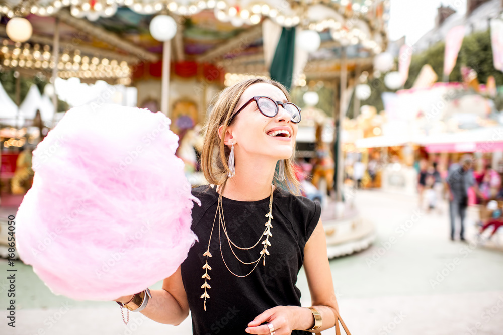 Fototapeta premium Young woman standing with pink cotton candy outdoors in front of the carrousel at the amusement park