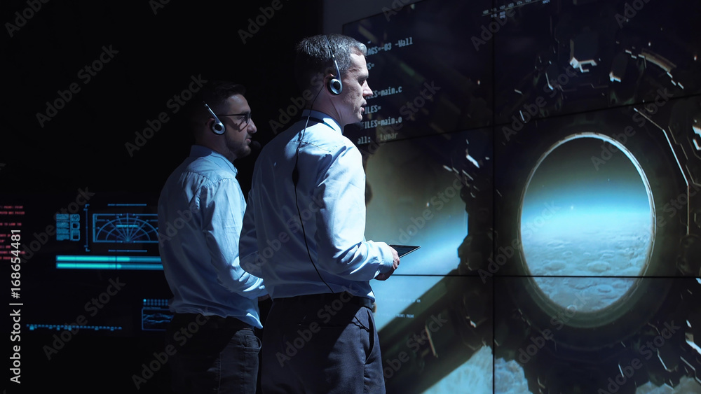 Back view of two supervisors standing at screen in space center and controlling landing on Moon. - obrazy, fototapety, plakaty 