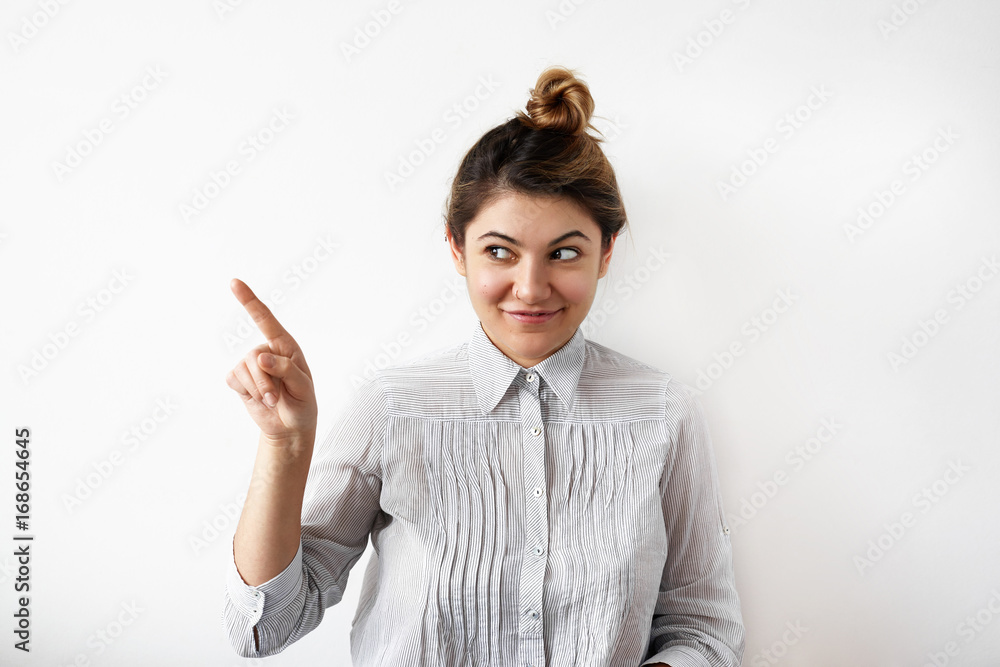 Happy positive young female secretary or manager with hair bun looking sideways with mysterious smile, pointing index finger at white wall with copy space for your promotional information on text Happy positive young female secretary or manager with hair bun looking sideways with mysterious smile, pointing index finger at white wall with copy space for your promotional information on text