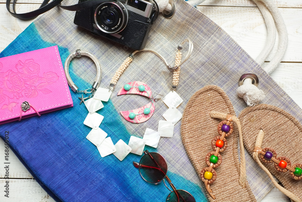 Flat lay of summer fashion with camera, slippers, sunglasses and other girl accessories on top of the bag on white wooden background Flat lay of summer fashion with camera, slippers, sunglasses and other girl accessories on top of the bag on white wooden background