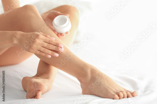 Beautiful young woman applying cream onto legs while sitting on bed at home. Epilation concept