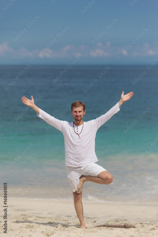 Handsome man in white clothes stand in yoga asana, tree pose, on sand beach. Yoga on the ocean in Bali. Handsome man in white clothes stand in yoga asana, tree pose, on sand beach. Yoga on the ocean in Bali.