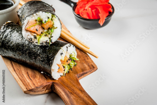 Trend hybrid food. Japanese, Asian cuisine. Sushi-burrito, sandwich with salmon, hayashi wakame, daikon, pickled ginger, red caviar. On a white marble table, with chopsticks and soy sauce. Copy space