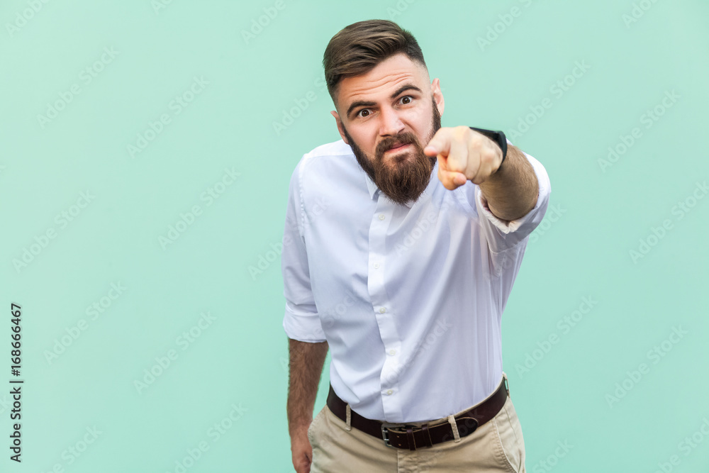 Angry bearded businessman with white shirt looking at camera and pointing finger to you. Studio shot isolated on light green background.