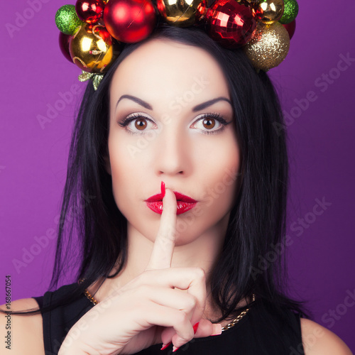 beautiful woman wearing a wreath made from Christmas decorations with a finger on her lips showing to keep silence