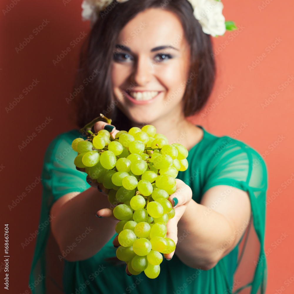 beautiful young woman wearing wreath and holding green grapes against red background beautiful young woman wearing wreath and holding green grapes against red background