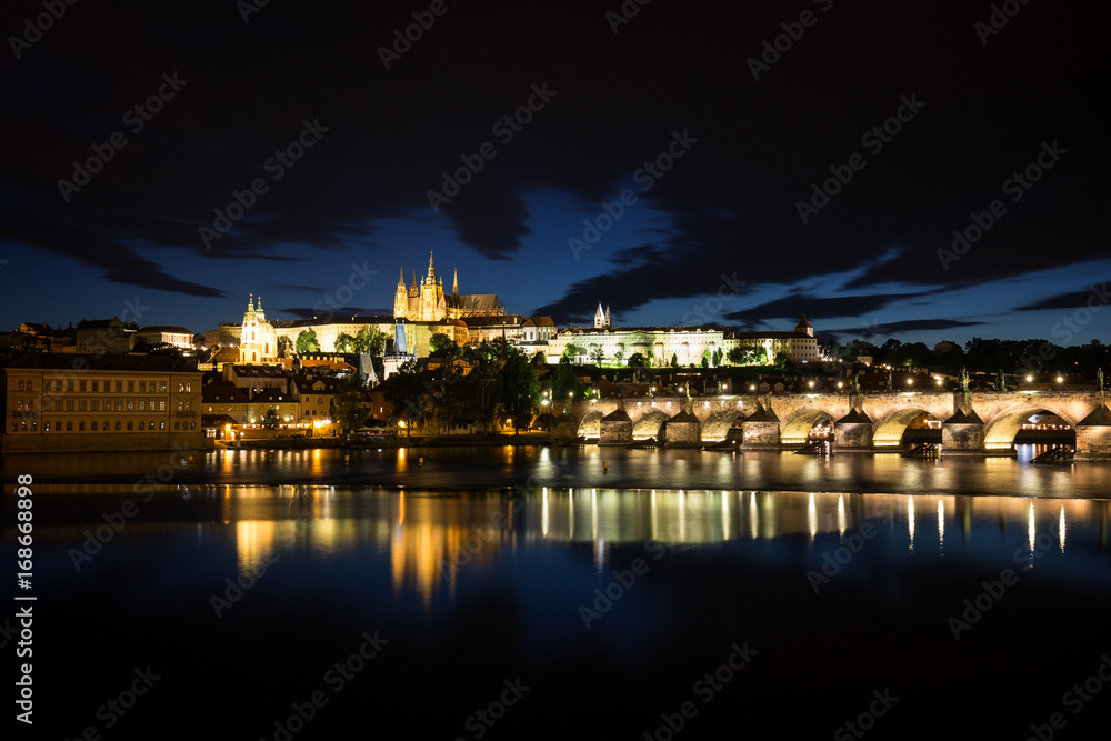 View of the lit Prague (Hradcany) Castle, Charles Bridge (Karluv most) and their reflections on the Vltava River in Prague, Czech Republic, at night.