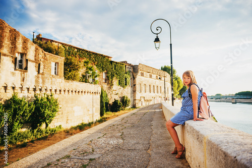 Canvas Print Kid girl tourist resting on quay of Arles, Provence, south of France, wearing blue gingham dress and pink backpack