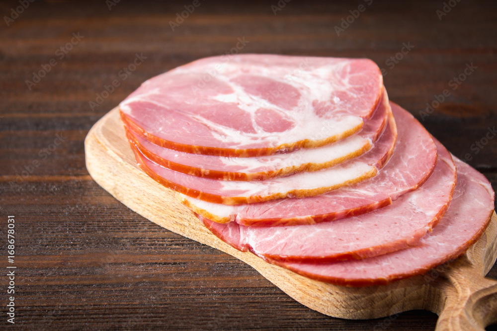 Sliced ham with fresh green lettuce leaves on a round cutting board. Meat products on a brown wooden table. Sliced ham with fresh green lettuce leaves on a round cutting board. Meat products on a brown wooden table.