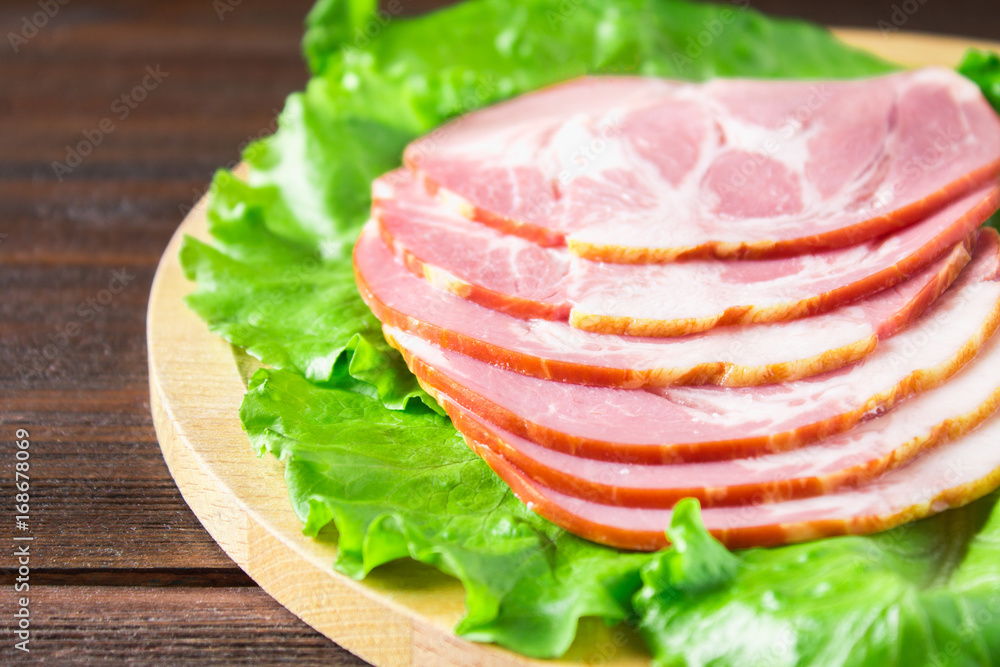 Sliced ham with fresh green lettuce leaves on a round cutting board. Meat products on a brown wooden table.