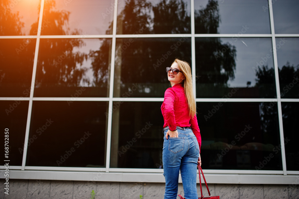 Portrait of a fabulous young woman in red blouse and jeans posing with her handbag and sunglasses outside the shopping mall on glass background.