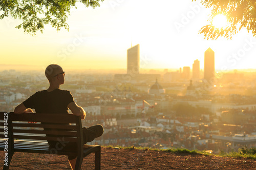 A man on a bench in a park, relaxing and  enjoying the summer sunrise over a city. Lyon, France.