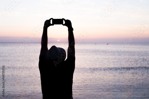 silhouette of a man holding a cellphone taking pictures outside during sunrise or sunset on the beach. Black light photo.