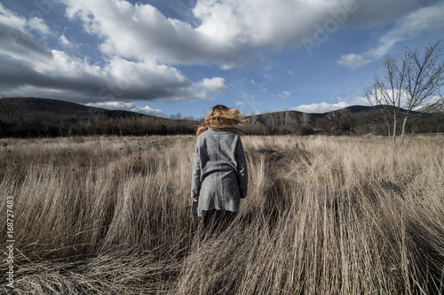 Back view of woman in gray coat standing in autumnal field on background of mountains. 