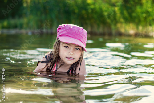 Fototapeta Naklejka Na Ścianę i Meble -  Happy little girl in pink cap having fun and swimming in lake water