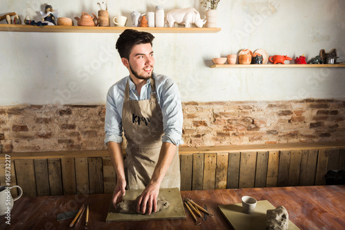 potter, workshop, ceramics art concept - standing young smiling brunette man with hands knead fireclay, male dressed in an apron, a ceramist with raw materials on wooden table with sculpting tools set