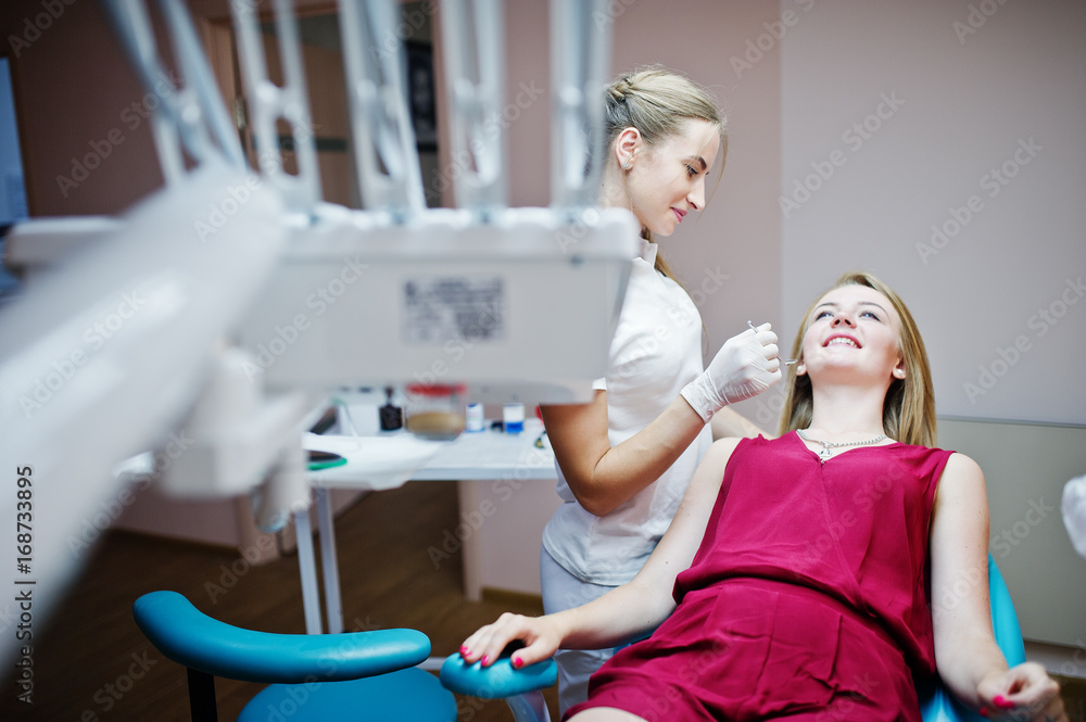 Obraz premium Attractive patient in red-violet dress laying on the dental chair while female dentist treating her teeth with special instruments.