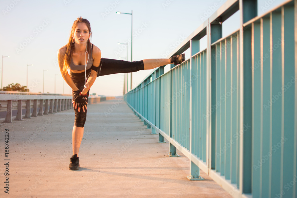 Young sporty woman stretching her legs leaning on bridge's fence outdoors.