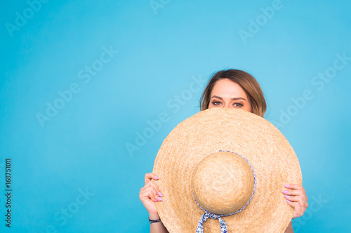 Beautiful young woman wears in summer dress and straw hat is laughing on blue background with copy space