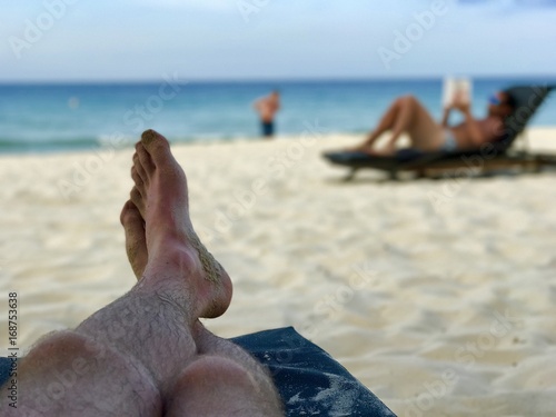 Legs on a beachchair and blurry man reading a book on the beach photo