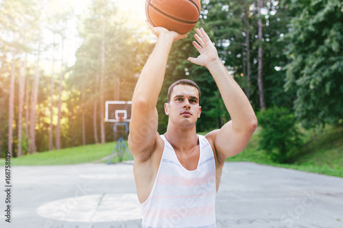 Young athletic man playing basketball in a beautiful park surrounded with trees.