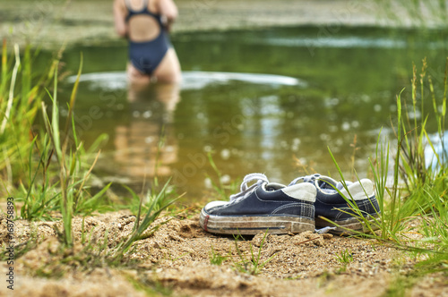 Old sneakers on the foreground and woman entering the pond's water on the blurred background      